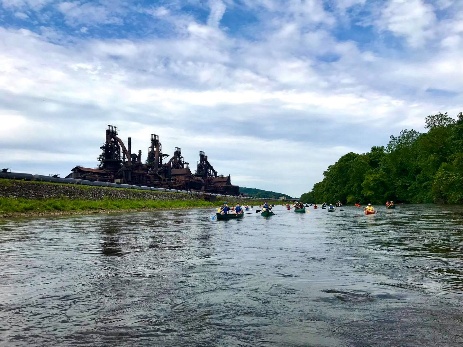 A group of people in canoes navigates a river under the backdrop of a large, industrial-looking factory structure.
AI-generated content may be incorrect.